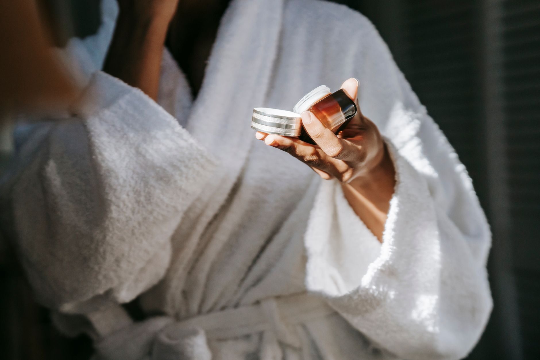 A hand in a soft robe holding a small amber glass jar in warm morning light